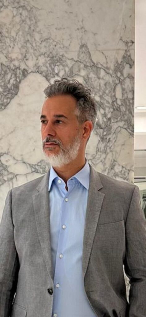 A man with grey hair and beard wearing a gray suit.